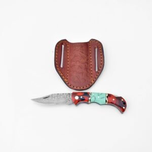 A folding knife with a Damascus blade and a red, black, and teal handle lies below a brown, embossed leather sheath