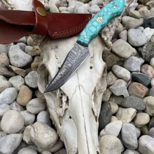 A Damascus steel knife with a turquoise and gold handle rests on a deer skull and antler, with a leather sheath