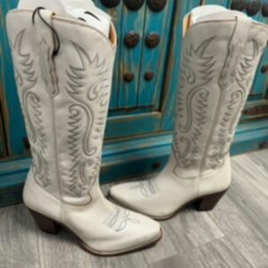 Two white cowboy boots with gray stitching and brown heels stand on a gray floor in front of a distressed turquoise cabinet