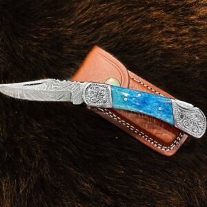An open folding knife with a wavy Damascus blade, vibrant blue handle, and engraved silver bolsters, on dark fur next to a brown leather sheath