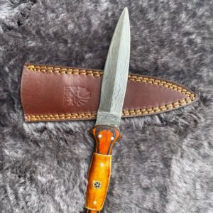 A knife with a wavy Damascus steel blade and an orange and tan handle, next to a brown leather sheath, lies on gray fur