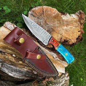 A Damascus steel knife with a patterned blade and a colorful handle lies next to its brown leather sheath on a log and grass