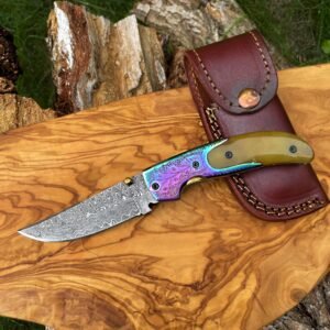 A Damascus steel folding knife with an iridescent handle and a brown leather sheath rest on a wooden board