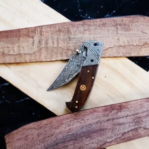 A folding knife with a Damascus steel blade and dark wood handle featuring an ornate emblem, resting on wood planks