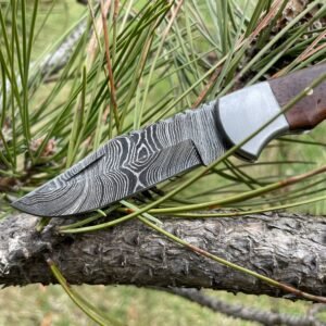 A knife with a wavy Damascus steel blade and a wood-and-bone handle lies on a pine branch