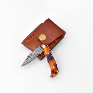 A folding knife with a Damascus steel blade and an amber-patterned handle rests next to a brown leather sheath with orange stitching