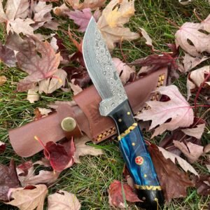 Damascus knife with a blue and black handle featuring gold and red accents, next to a brown leather sheath on autumn leaves and grass