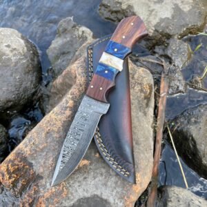 A Damascus steel knife with a segmented wood, blue, and off-white handle rests on a wet rock with its leather sheath next to water