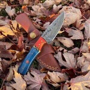 A knife with a swirling patterned blade and a red-orange to blue handle is partially out of a brown leather sheath on autumn leaves and grass