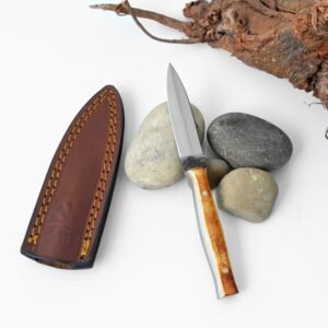 A knife with a two-toned handle and brown leather sheath rests among smooth stones and gnarled wood on a white background