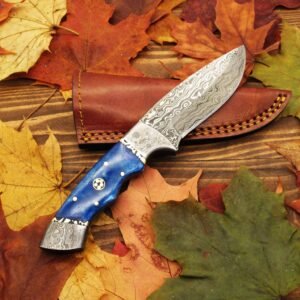 A knife with a patterned Damascus steel blade and a blue handle with brass pins rests on a wooden surface surrounded by colorful autumn leaves