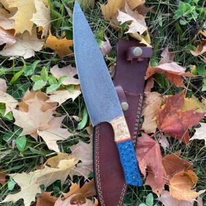 A knife with a Damascus patterned blade and a textured blue and wood handle is in a brown leather sheath on autumn leaves and grass