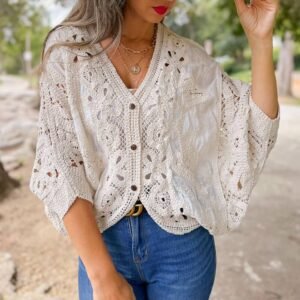 A person in a white wide-brim hat, cream crochet top with brown buttons, blue jeans, and layered gold necklaces, outdoors
