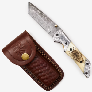 A folding knife with a Damascus steel blade and an ornate handle depicting an eagle and a wolf lies next to a textured leather sheath