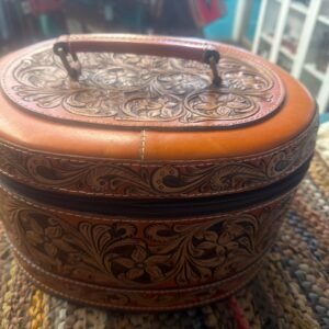 A round, light brown leather case with intricate floral tooling, a top handle, and a zipper, resting on a woven mat