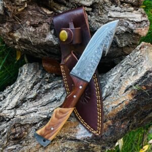 A Damascus steel knife with a two-toned wooden handle in a brown leather sheath with yellow stitching on a tree log