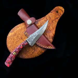A custom knife with a patterned Damascus blade and a red marbled handle lies on a burlwood board, next to its brown leather sheath