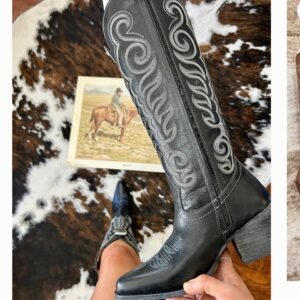 A person holds up a black cowboy boot with intricate white scroll stitching, with another boot and a cowhide rug in the background