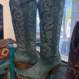A pair of dark teal cowboy boots with intricate white swirling embroidery and wooden heels is displayed in a window