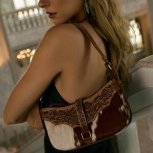 A woman wearing a dark dress holds a brown leather shoulder bag with white and brown cowhide and intricate tooling