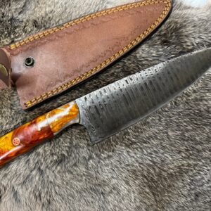 A Damascus steel knife with a colorful orange and red resin handle lies next to its brown leather sheath on gray fur