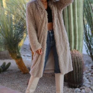 A woman in a long beige faux fur coat, blue jeans, and white boots stands in a desert garden with cacti