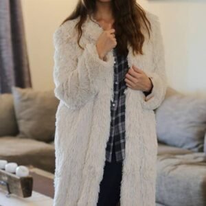 A woman with long dark hair wears a light beige faux fur coat over a dark plaid shirt, standing indoors