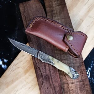 A folding knife with a Damascus blade, bone handle, and engraved metal details next to a brown leather sheath on wood and dark marble