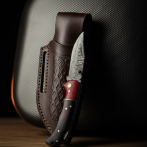 A Damascus steel knife with a red and dark wood handle rests next to an embossed brown leather sheath