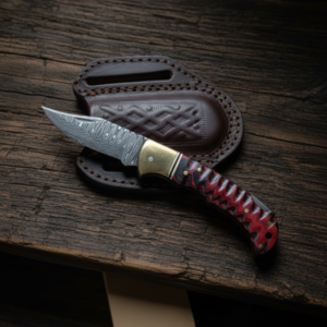 A folding knife with a Damascus blade and a red and black handle rests on a brown leather sheath on a wooden surface