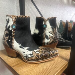 A pair of black and white cowhide cowboy boots with floral tooling and turquoise accents on a wooden shelf