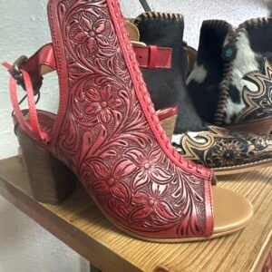 A red open-toe leather shoe with intricate floral carvings sits on a wooden shelf, with cowboy boots in the background