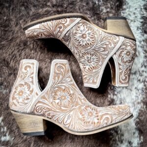 Two white leather boots with intricate brown floral tooling, one an ankle boot and one a high-heeled mule, lie on a fur rug