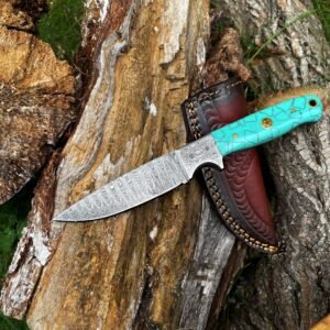 A Damascus steel knife with a turquoise handle rests on a weathered log beside a dark leather sheath