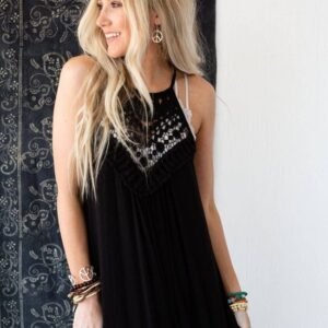 A smiling blonde woman wears a black maxi dress with a white crochet detail, peace sign earrings, and stacked bracelets