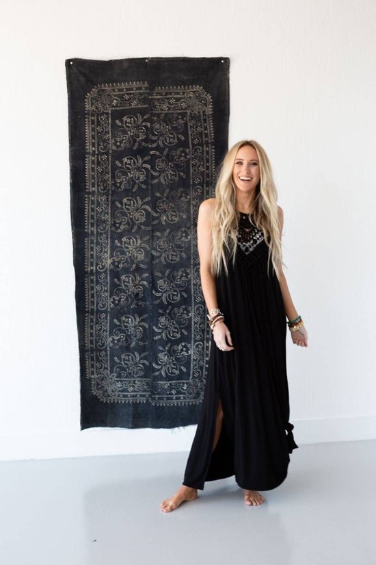 Smiling woman in a black maxi dress with a detailed halter neckline, standing barefoot by a dark patterned tapestry