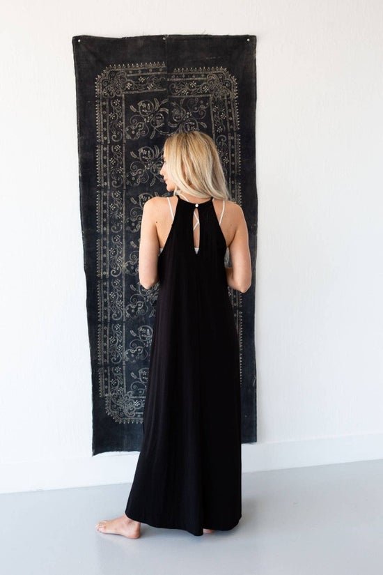 A blonde woman in a black halter maxi dress with a keyhole back stands facing away, looking over her left shoulder, in front of a dark patterned tapestry