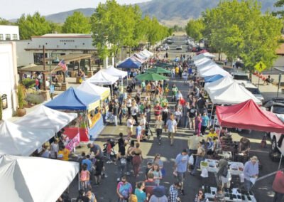 A lively street market packed with people browsing stalls under white and colorful tents, with trees and mountains in the background