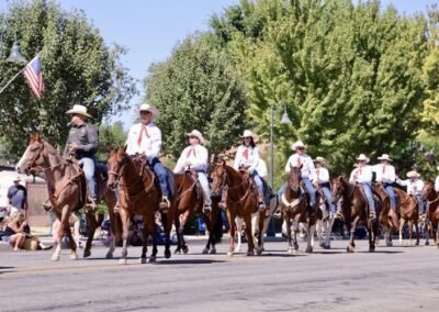 A group of people in cowboy hats and white shirts ride horses in a parade, led by a man in uniform