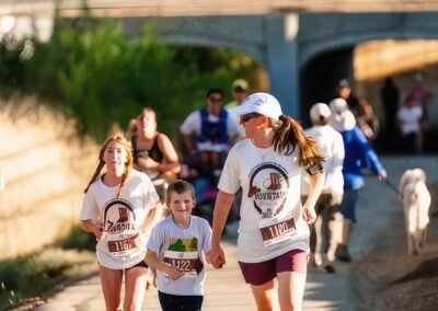 A woman, a boy, and a girl with race bibs run on a paved path, the woman and boy holding hands, with other runners in the background